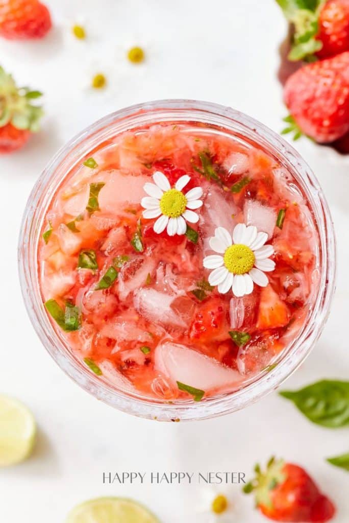 A refreshing strawberry basil cocktail with crushed strawberries, ice, and garnished with two small white flowers. Surrounded by fresh strawberries, lime slices, and basil leaves on a light background.