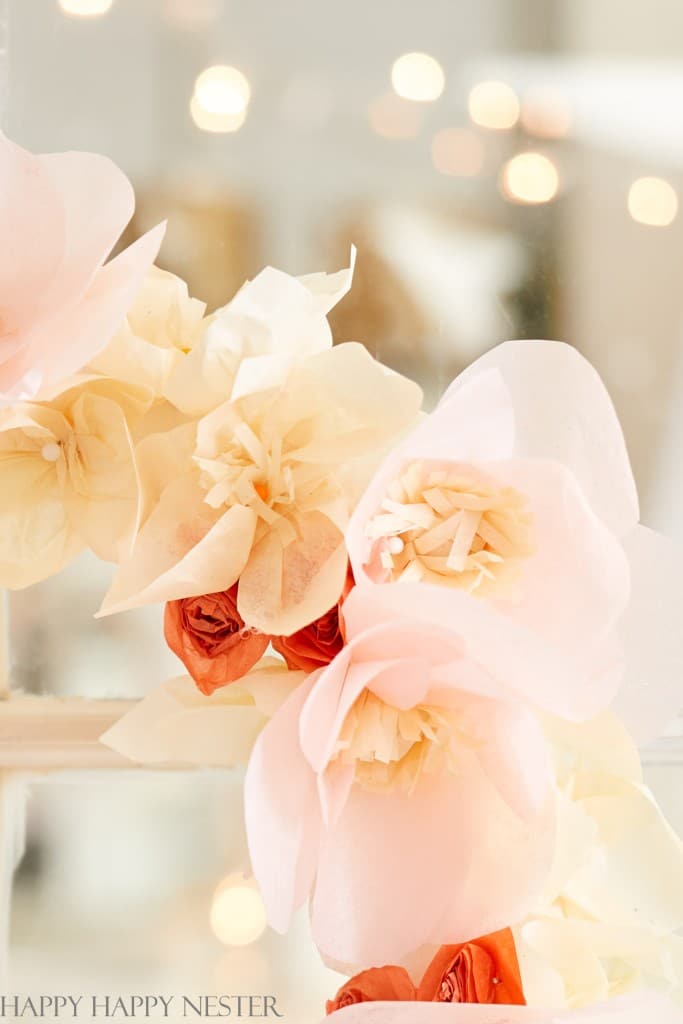 Close-up of delicate, pastel-colored paper flowers crafted in soft pink and cream, with small red blossoms interspersed. These easy flower paper crafts are set against a blurred background with warm bokeh lights, creating a dreamy, romantic atmosphere.