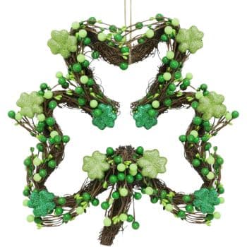 A star-shaped wreath made of intertwined twigs is adorned with green shamrock decorations and green berries. It hangs against a plain white background.