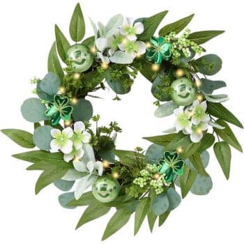 A decorative wreath featuring green leaves, white and green flowers, and green ornaments. Shamrock-shaped embellishments and small yellow lights are woven through the foliage.