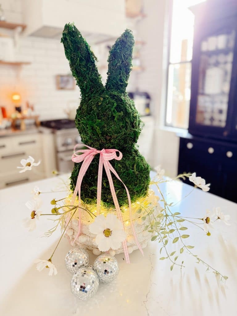 A moss-covered bunny sculpture with a pink ribbon sits on a table decorated with white flowers and small disco balls. Warm lights wrap around the sculpture. The background features a white kitchen with natural lighting.