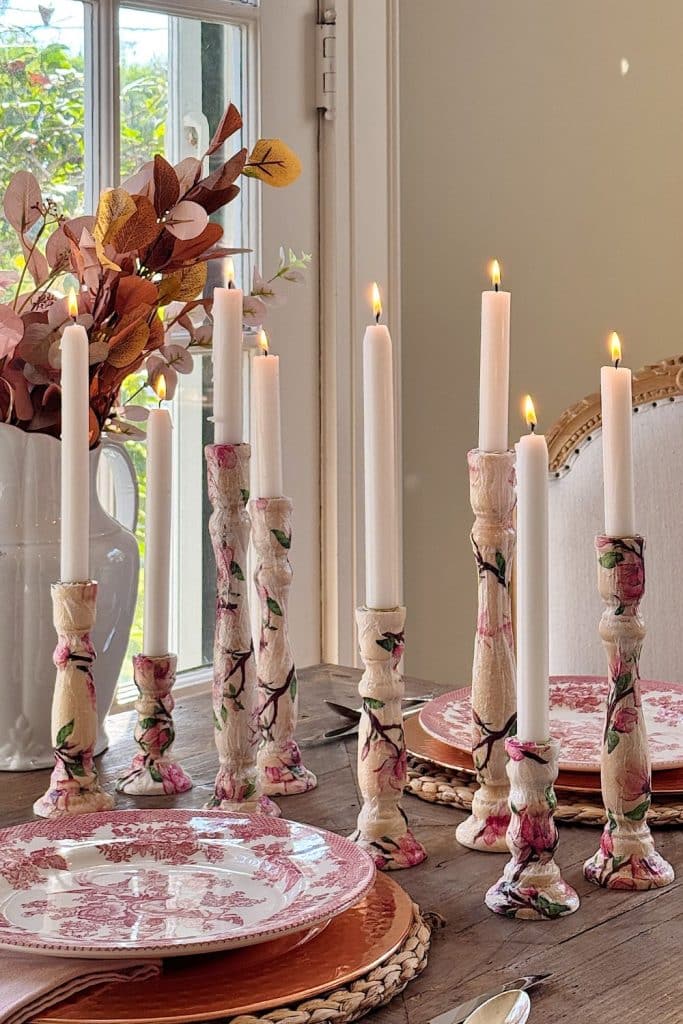 Dining table set with tall, lit white candles in decorative holders adorned with pink floral patterns. The table features pink and white patterned plates, a woven placemat, and a large white vase with autumn-colored foliage, beside a window.