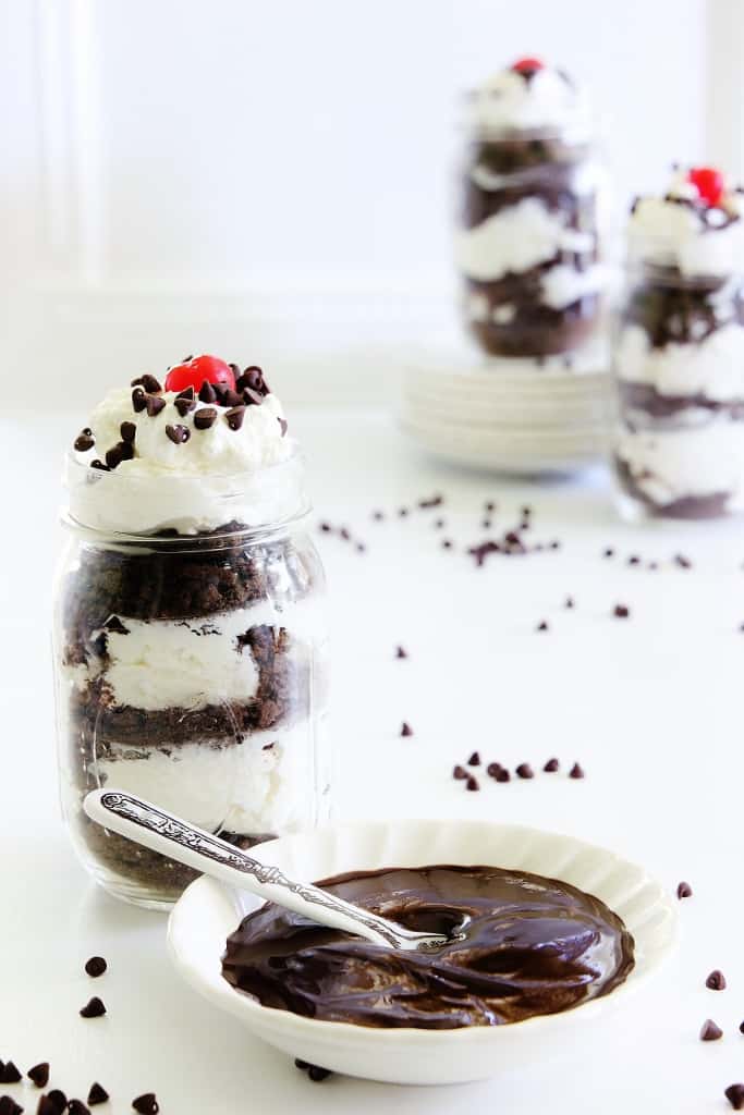 A glass jar brimming with layers of chocolate cake and whipped cream, crowned with a cherry and chocolate chips, captures the essence of the best spring cakes. Nearby, a small bowl of chocolate sauce awaits with a spoon. In the background, more jars and scattered chocolate chips complete the scene.