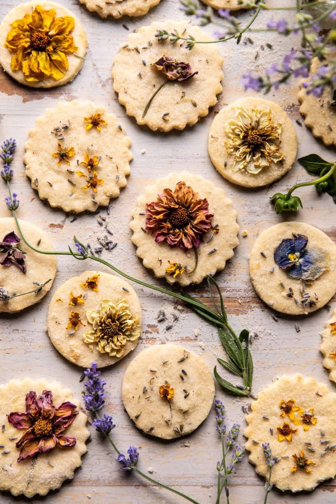 A selection of favorite spring cookies topped with colorful edible flowers, like marigolds and violets, are arranged on a light wooden surface. Sprigs of lavender and other herbs are scattered around, adding a rustic feel.