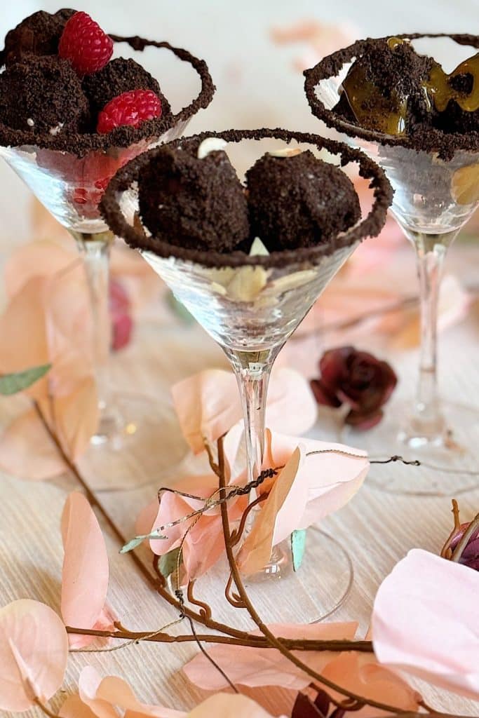 Three martini glasses filled with chocolate truffles, garnished with chocolate crumbs. One glass has raspberries, another has kiwi slices, and the third features a hint of Oreo. Surrounded by pink floral decor on a light wooden surface.