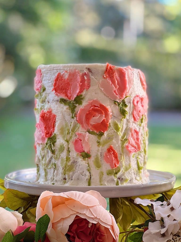 A tall, round cake on a white stand is adorned with pink and green floral buttercream designs, exemplifying one of the best spring cakes. The background is a blurred, lush green outdoor scene with soft focus on surrounding flowers.