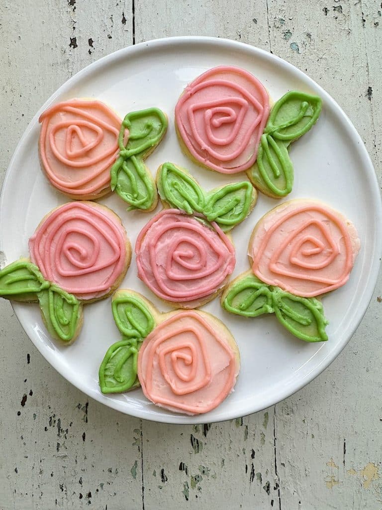 A white plate holds six of my favorite spring cookies, each decorated with pink icing swirls forming roses and adorned with green icing leaves. The background is a rustic, pale wooden surface.