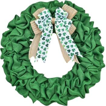 A green fabric wreath with ruffled texture features a large bow at the top. The bow consists of a burlap layer beneath a white ribbon decorated with green shamrocks.