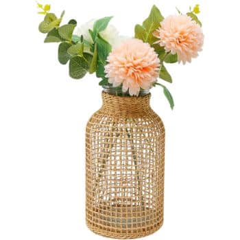 A decorative vase with a woven wicker exterior holds an arrangement of light pink flowers and green leaves. The flowers have a round, fluffy appearance, resembling dahlias or chrysanthemums.