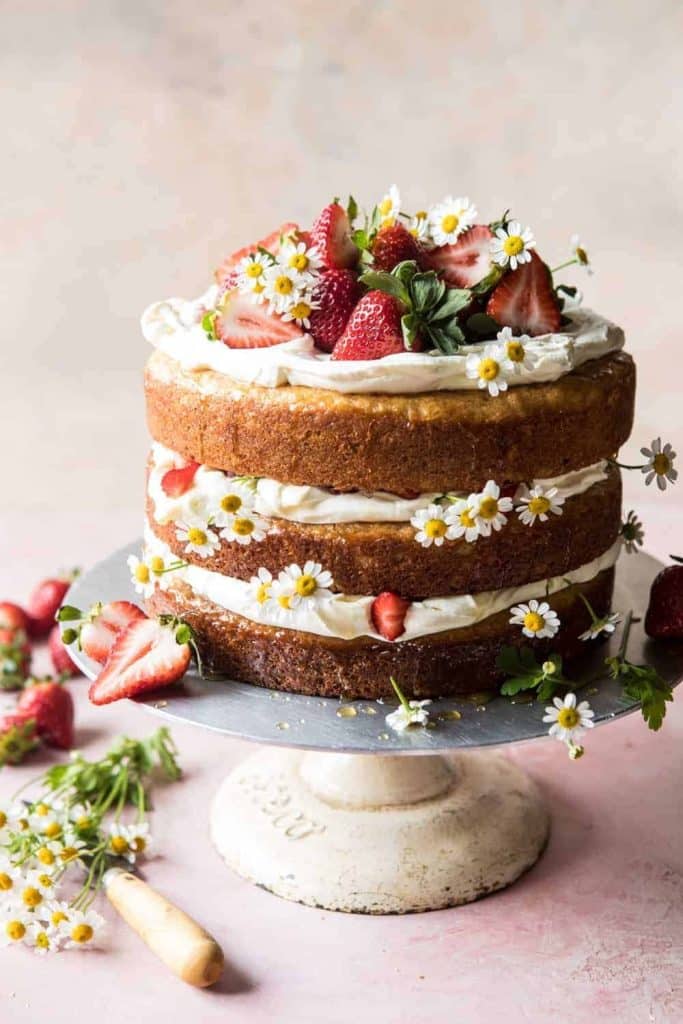 This three-layer naked cake, one of the best spring cakes, features cream and strawberries between layers. It's elegantly topped with fresh strawberries and small white flowers, all presented on a white cake stand against a soft pink background.