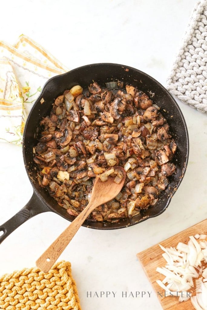 A black skillet brimming with cooked mushrooms and diced onions sits invitingly with a wooden spoon, hinting at a delicious homemade sourdough stuffing recipe. Nearby, a yellow woven potholder rests alongside chopped onions on a wooden cutting board.