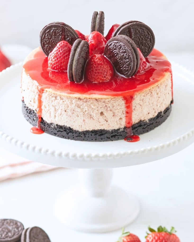 Experience one of the best spring cakes—a cheesecake on a white stand with a chocolate cookie crust, adorned with fresh strawberries and chocolate sandwich cookies, and drizzled with red syrup.