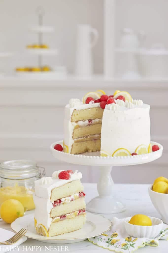 A layered lemon cake with white frosting on a cake stand, garnished with lemon slices and raspberries—one of the best spring cakes. A slice is cut out and placed on a plate beside a jar of lemon curd, a few lemons, and a floral napkin.