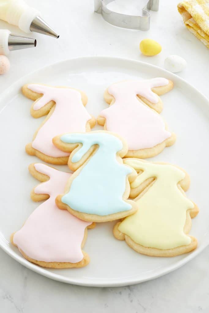 A plate of favorite spring cookies, shaped like bunnies and adorned with pastel icing in pink, blue, and yellow hues. Cookie cutters and baking tools are scattered on the light countertop in the background.