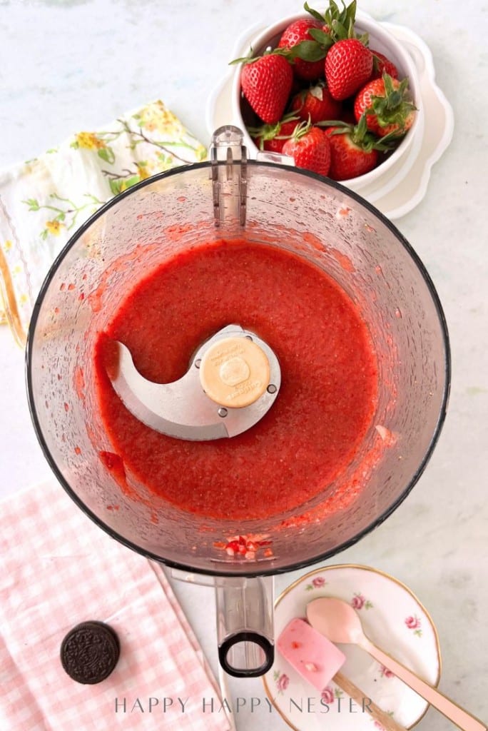 A top view of a food processor containing blended strawberry puree. Nearby are fresh strawberries in a bowl, a floral plate with a pink spatula, a pink checkered cloth, and an Oreo cookie. The background has a light, textured surface.