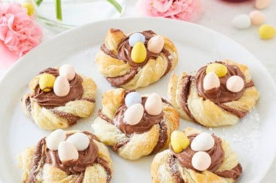A plate with five twisted pastries topped with chocolate spread and small colorful candy eggs. Surrounding the plate are pink flowers, a lit candle, and scattered candy eggs on a table with a checkered cloth.