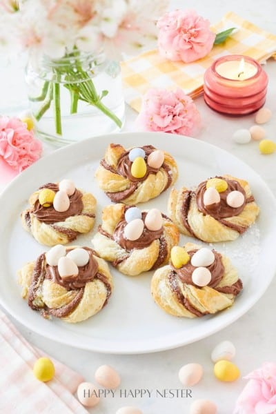 A plate with five twisted pastries topped with chocolate spread and small colorful candy eggs. Surrounding the plate are pink flowers, a lit candle, and scattered candy eggs on a table with a checkered cloth.