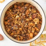 A pot of savory stuffing filled with pieces of homemade sourdough, mushrooms, and herbs. The dish is browned on top, with visible chunks of garlic and onions. Surrounding the pot are some loose croutons and a small jar labeled "Herbes de Provence.