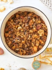 A pot of savory stuffing filled with pieces of homemade sourdough, mushrooms, and herbs. The dish is browned on top, with visible chunks of garlic and onions. Surrounding the pot are some loose croutons and a small jar labeled "Herbes de Provence.