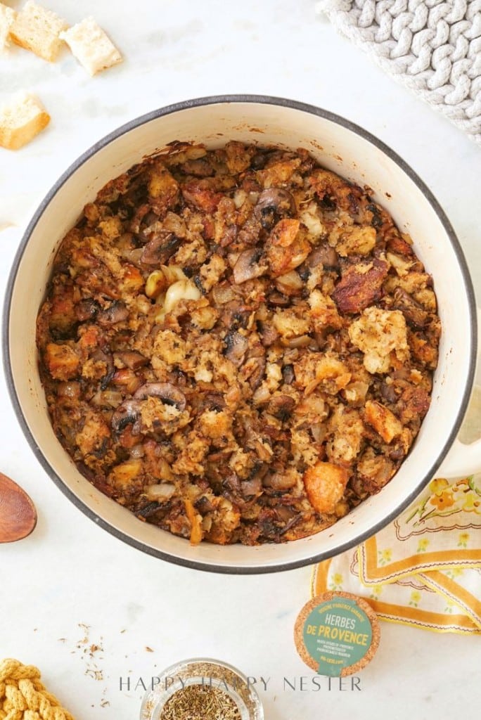 A pot of savory stuffing filled with pieces of homemade sourdough, mushrooms, and herbs. The dish is browned on top, with visible chunks of garlic and onions. Surrounding the pot are some loose croutons and a small jar labeled "Herbes de Provence.