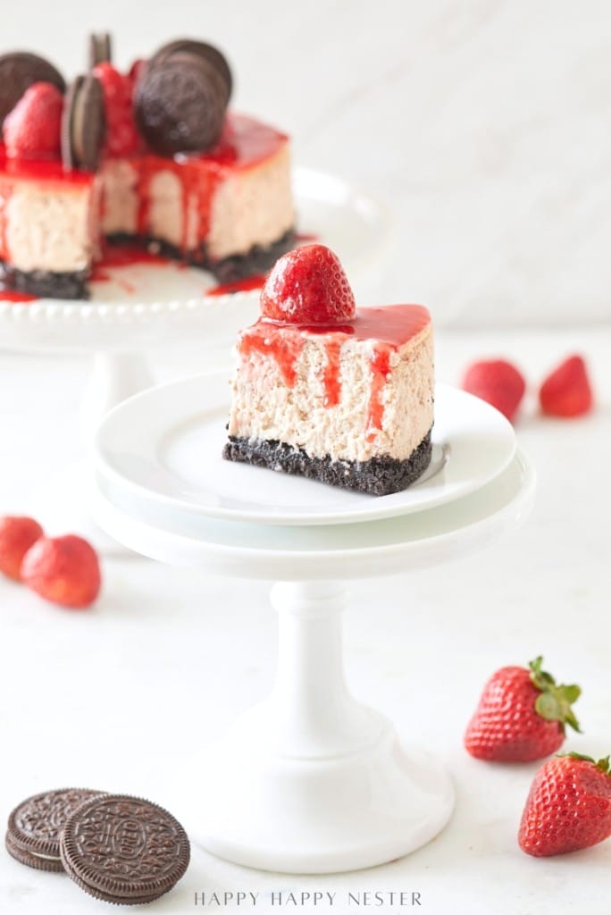 A slice of Oreo Strawberry Cheesecake is showcased on a white cake stand, crowned with a fresh strawberry and drizzled with strawberry sauce. In the background, discover the tempting Oreo Strawberry Cheesecake Recipe featuring a whole cheesecake adorned similarly, surrounded by Oreo cookies and scattered strawberries.