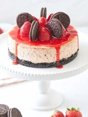 Decadent strawberry cheesecake topped with whole chocolate sandwich cookies and fresh strawberries, drizzled with a vibrant red sauce, sits on a white cake stand against a soft background.