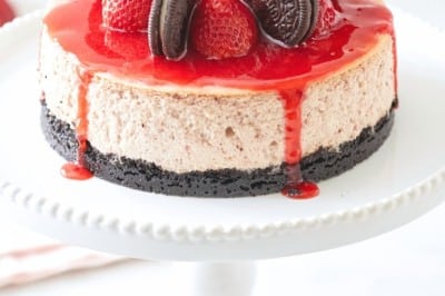 Decadent strawberry cheesecake topped with whole chocolate sandwich cookies and fresh strawberries, drizzled with a vibrant red sauce, sits on a white cake stand against a soft background.
