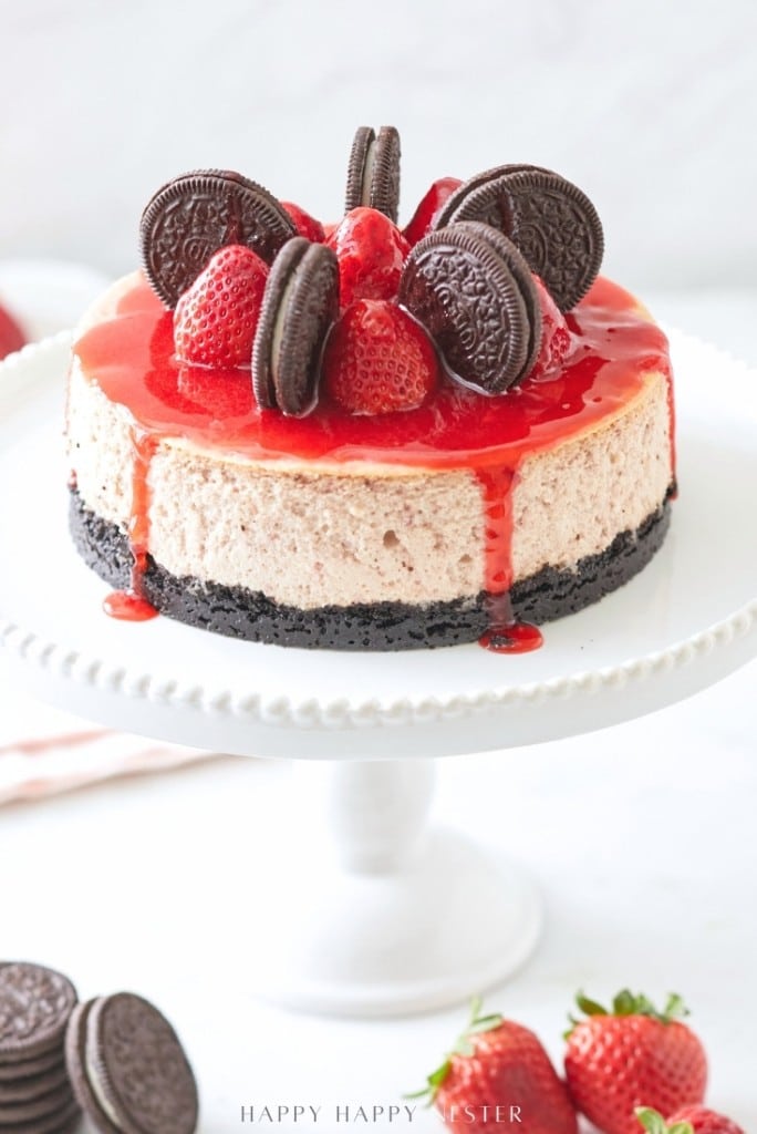 Decadent strawberry cheesecake topped with whole chocolate sandwich cookies and fresh strawberries, drizzled with a vibrant red sauce, sits on a white cake stand against a soft background.