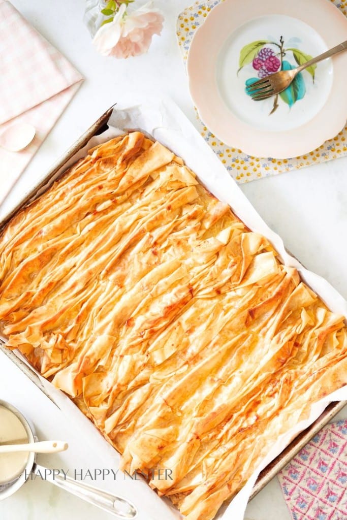 A tray of crispy, golden-brown phyllo pastry, one of the best spring cakes, sits on a table with a vintage fork and a floral plate nearby. Pink and white flowers and a pastel napkin complete the elegant setting.