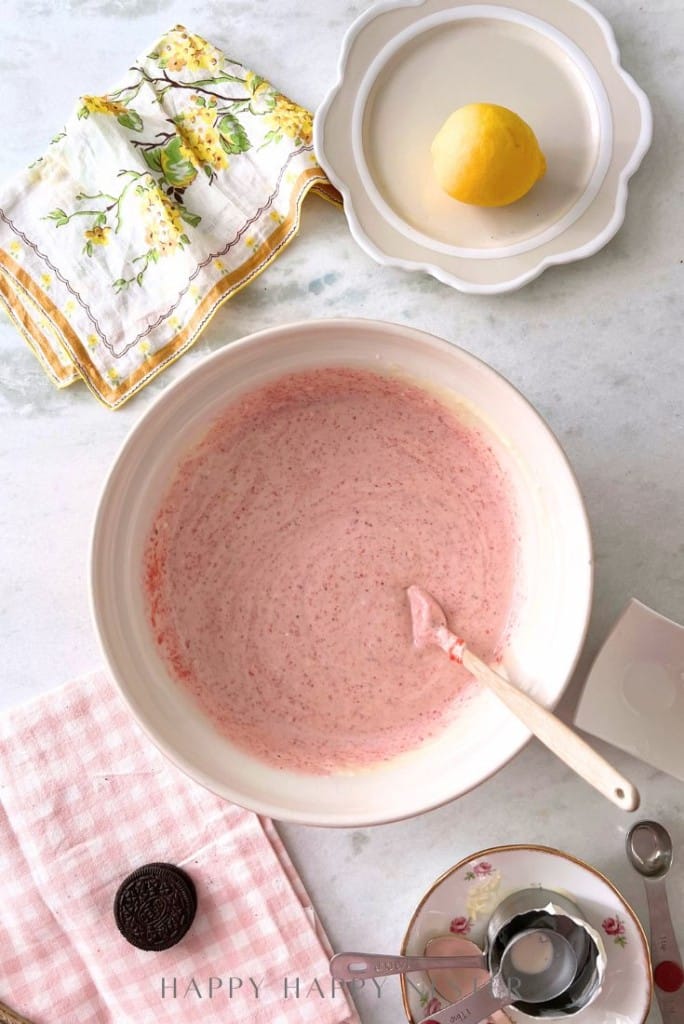 A mixing bowl with pink batter and a spatula sits on a marble countertop. Next to it are a lemon, patterned napkin, Oreo cookie, and a floral plate. A pink checkered cloth is beneath.