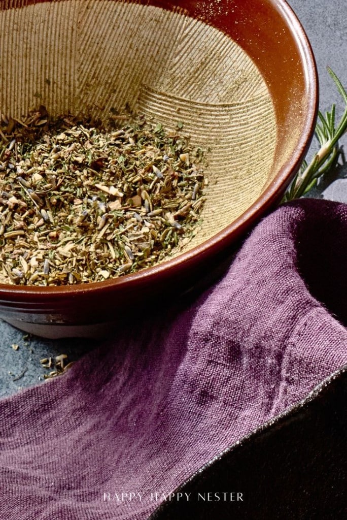 A textured brown bowl contains mixed dried herbs, placed on a dark surface. A sprig of rosemary lies nearby. A dark purple cloth partially covers the bottom of the image.