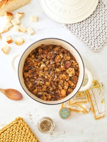 A white pot filled with homemade sourdough stuffing sits on a marble countertop. Surrounding it are bread pieces, a spoon, a jar of herbs, colorful napkins, and two woven pot holders. A loaf of bread and "Happy Happy Nester" text complete the cozy scene.