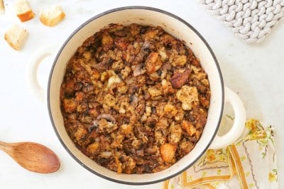 A white pot filled with homemade sourdough stuffing sits on a marble countertop. Surrounding it are bread pieces, a spoon, a jar of herbs, colorful napkins, and two woven pot holders. A loaf of bread and "Happy Happy Nester" text complete the cozy scene.
