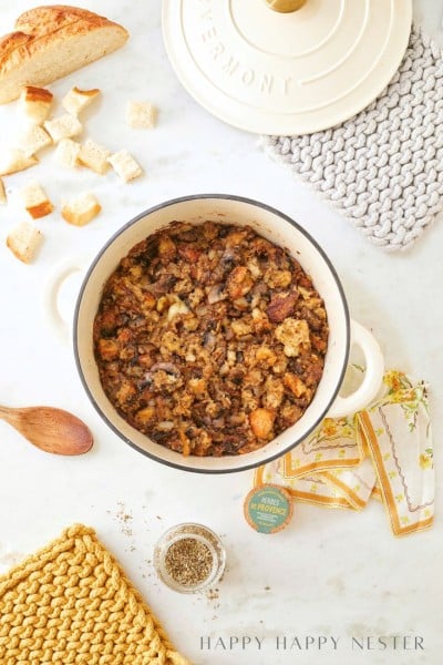 A white pot filled with homemade sourdough stuffing sits on a marble countertop. Surrounding it are bread pieces, a spoon, a jar of herbs, colorful napkins, and two woven pot holders. A loaf of bread and "Happy Happy Nester" text complete the cozy scene.