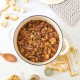 A white pot filled with homemade sourdough stuffing sits on a marble countertop. Surrounding it are bread pieces, a spoon, a jar of herbs, colorful napkins, and two woven pot holders. A loaf of bread and "Happy Happy Nester" text complete the cozy scene.