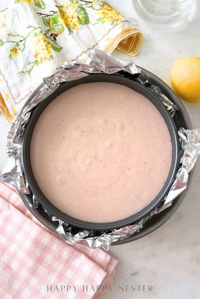 Cake batter in a round pan lined with foil, placed on a marble surface. A pink checkered cloth, lemon, and floral cloth are nearby. The batter is light pink, suggesting a fruity flavor.