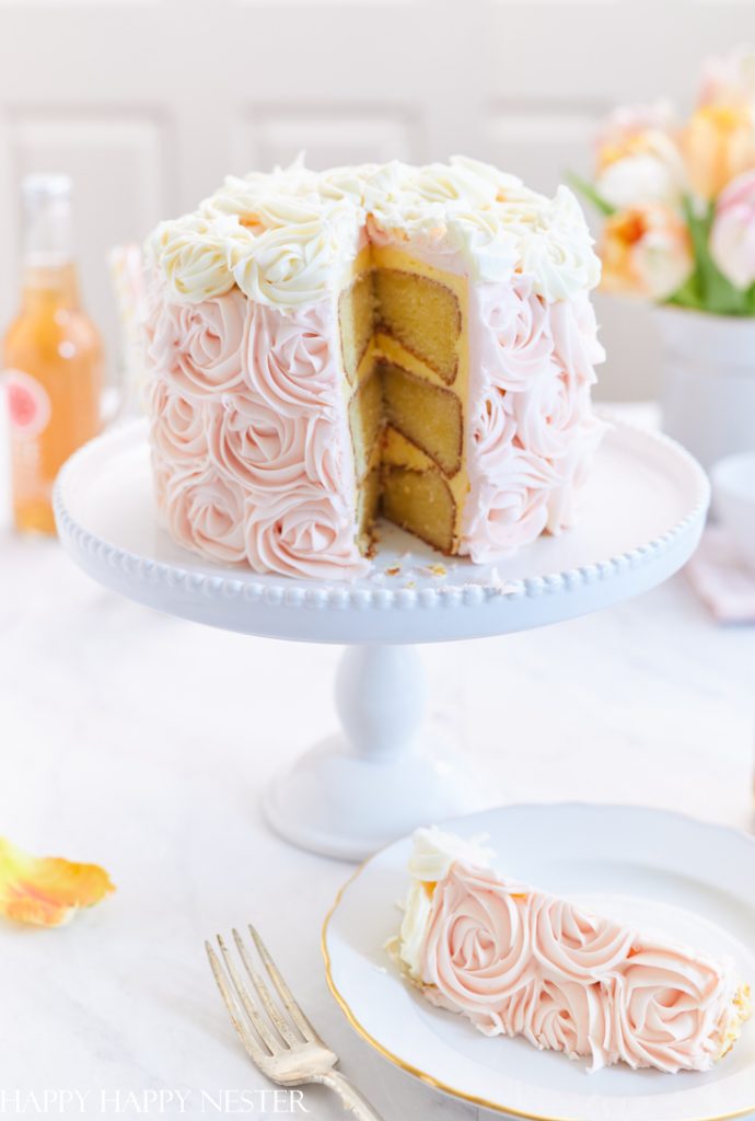 A slice of cake with delightful pink and white rosette frosting sits on a white plate beside a cake stand holding the rest of one of the best spring cakes. The interior reveals three layers, while the background features a bottle and a vase brimming with flowers.