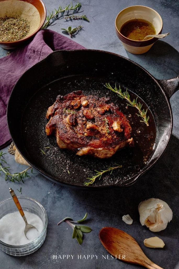 Learn how to prepare perfectly cooked steak garnished with herbs and garlic in a black skillet on a blue surface. Nearby, you'll find a small bowl of oil, fragrant herbs, salt, a wooden spoon, garlic cloves, and an elegant purple cloth.