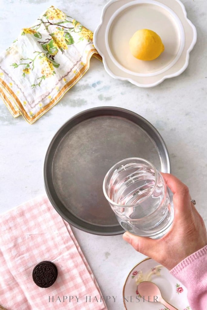 A hand holding a glass of water above a round metal pan. A lemon on a plate, a decorative napkin, a pink checkered cloth, and a chocolate cookie on a saucer are arranged on a light countertop.