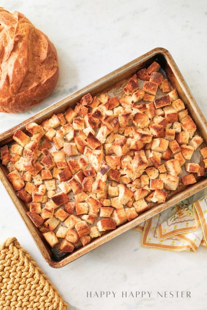 A baking tray filled with golden-brown croutons sits on a marble surface. To the side, there's a round loaf of bread, a set of decorative napkins, and a woven coaster. Text at the bottom reads "Happy Happy Nester.