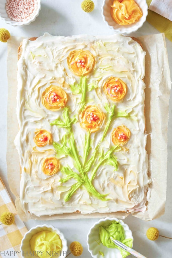 A rectangular cake frosted with creamy white icing features a bouquet of yellow and orange flower designs. Green icing forms stems and leaves. Surrounding the cake are small bowls of different colored favorite spring cookies and decorations on a checkered cloth.