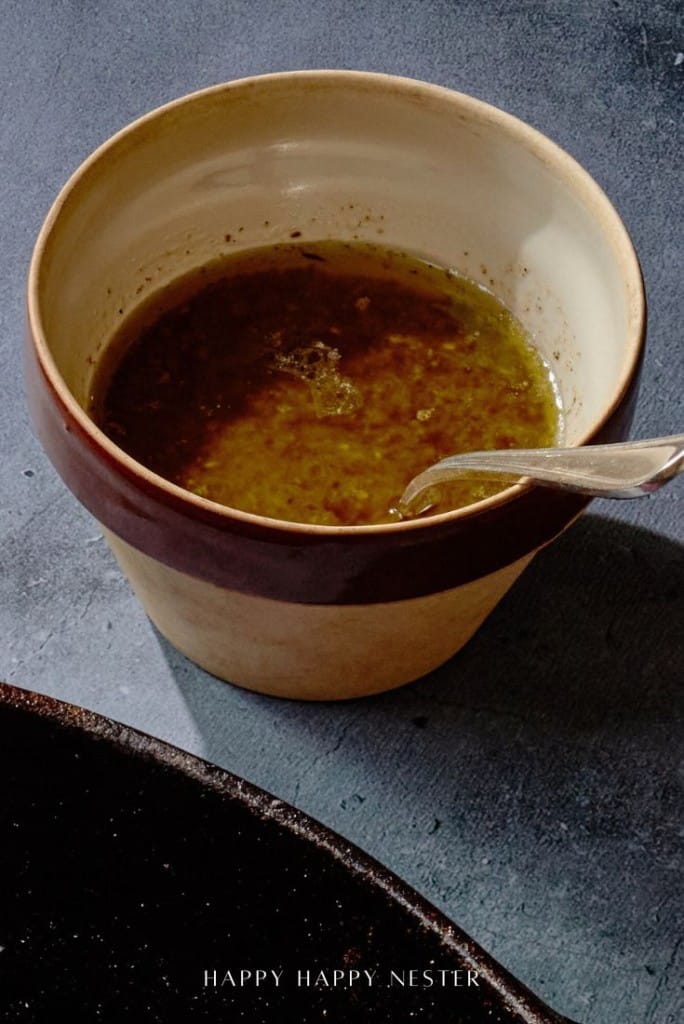 A ceramic cup filled with a brownish-green liquid, likely sauce or dressing for your tender, juicy steak, sits on a gray surface. A spoon rests inside the cup. The cup has brown and cream bands, and the base of a pan is visible in the corner.