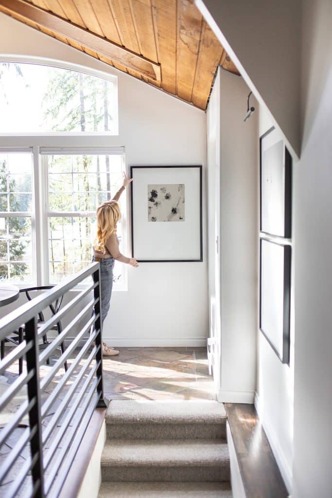 A person with long hair stands at the top of a narrow staircase, adjusting a framed artwork on a white wall. Sunlight streams through large windows, illuminating the wooden ceiling and black metal railing.