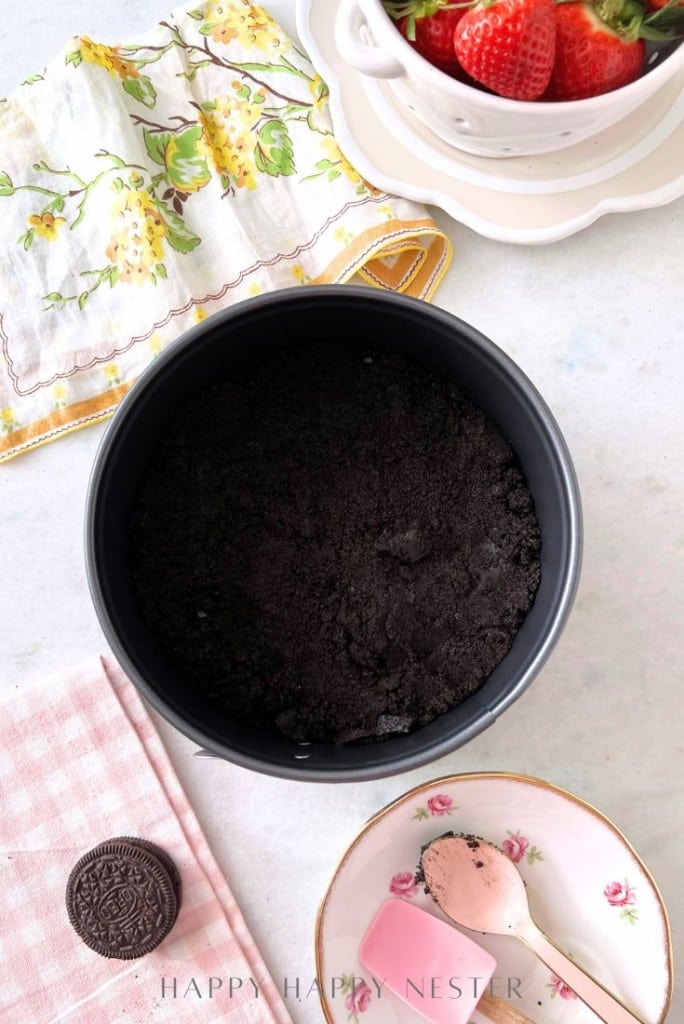 A round baking pan filled with crumbled chocolate crust sits on a marble surface. Nearby are a napkin with lemon prints, fresh strawberries in a strainer, a pink gingham cloth, an Oreo cookie, and a floral dish with a pink scoop.