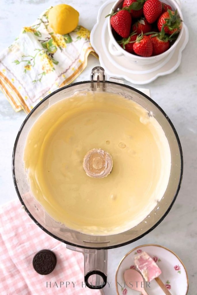 A food processor filled with creamy, smooth cheesecake batter. Nearby are fresh strawberries in a bowl, a lemon, a floral napkin, a pink checkered cloth, and plates with a slice of cheesecake and an Oreo cookie.