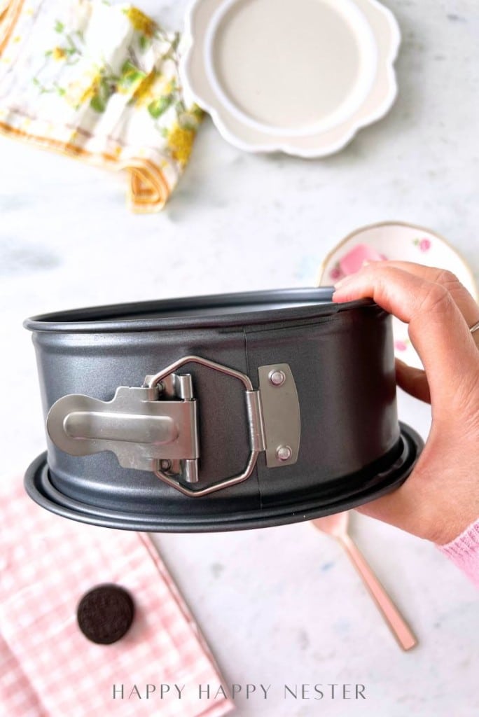 A hand holds a metal springform pan above a table. Nearby are a checkered cloth, a pink-handled spoon, a white plate, and a patterned napkin, setting the scene to create an Oreo Strawberry Cheesecake recipe alongside chocolate cookies.