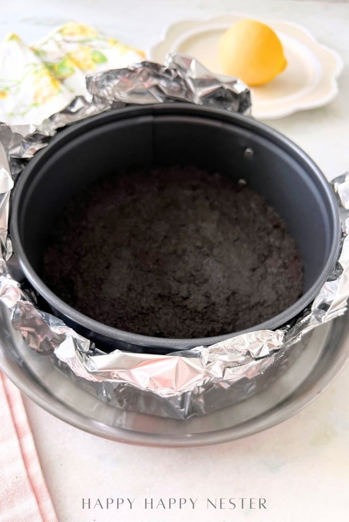 A round, foil-wrapped baking pan with a dark crust at the bottom sits on a metal tray. A lemon rests on a white plate in the background, next to a floral-patterned cloth. The text "HAPPY HAPPY NESTER" is visible at the bottom, hinting at an Oreo Strawberry Cheesecake recipe.