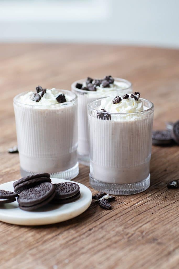 Three glasses of creamy milkshakes topped with whipped cream and crushed cookies sit on a wooden table. Nearby, a small plate holds whole and broken Oreo cookies, adding an irresistible touch to the indulgent scene.