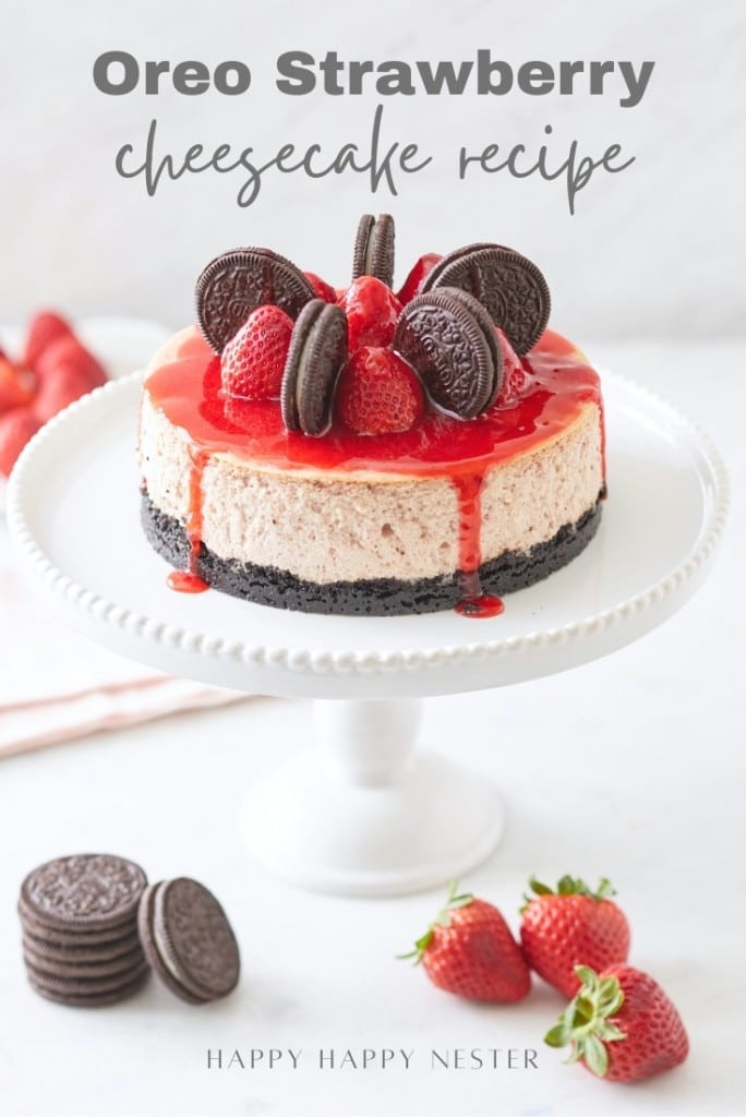 A strawberry cheesecake topped with whole strawberries, Oreo cookies, and a drizzle of strawberry sauce sits elegantly on a white stand. Fresh strawberries and Oreos are scattered at the base. Above the cake floats "Oreo Strawberry Cheesecake Recipe.