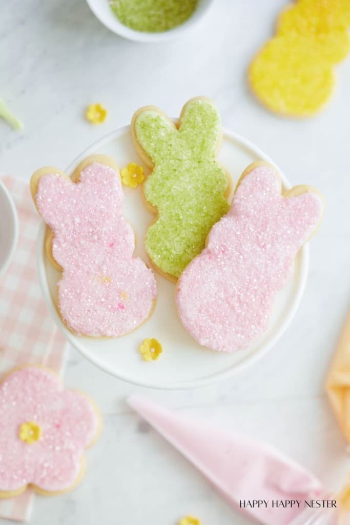 Three bunny-shaped sugar cookies, your favorite spring treats, rest on a white platter. Two are pink, one is green, all sprinkled with sugar. Nearby, a yellow flower-shaped cookie and small decorative flowers adorn the table's pink checkered cloth.